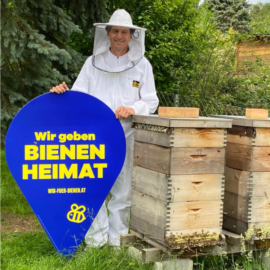 Höllinger Bienen - Jetzt online bei Höllinger bestellen! Gerhard Höllinger im Imker-Gewand mit einem Schild das "Wir geben Bienen Heimat" sagt vor zwei Bienenstöcken in freier Natir