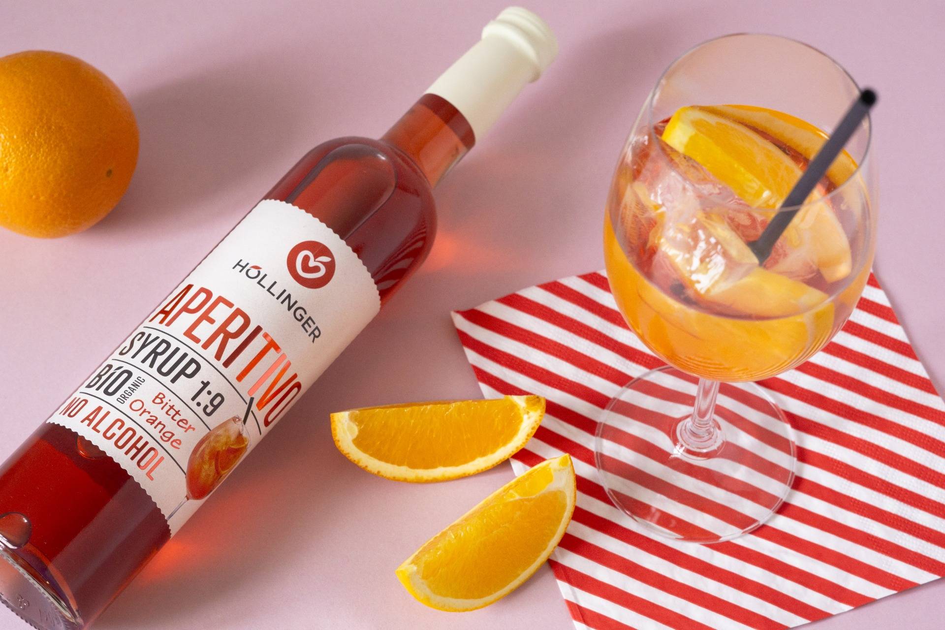 Hollinger Aperitivo Sirupflasche mit Glas Aperitivo Spritz und frischen Orangenscheiben auf rosa Hintergrund.