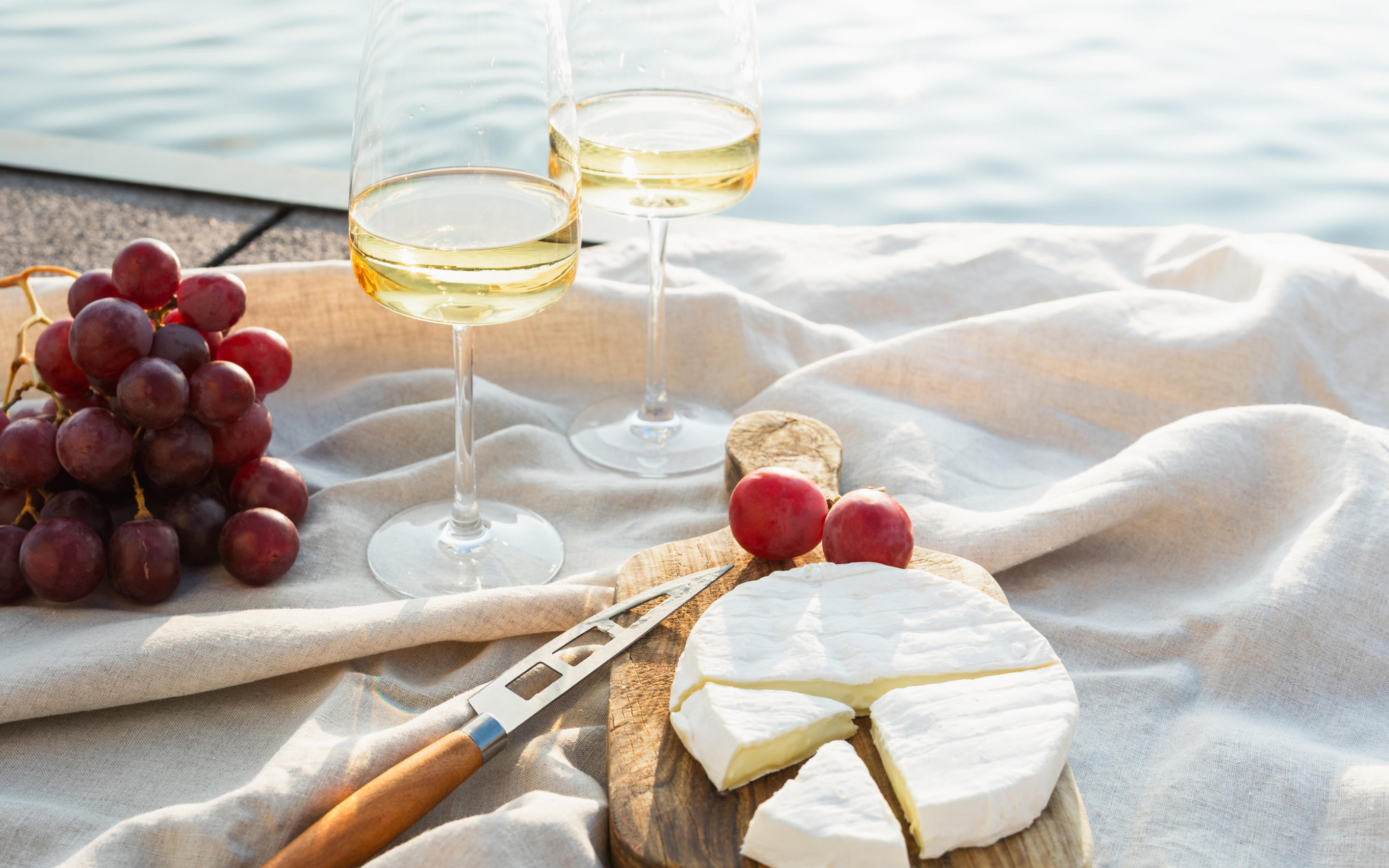 Zwei Gläser Secco mit Trauben und Käse auf einem Tuch am Wasser