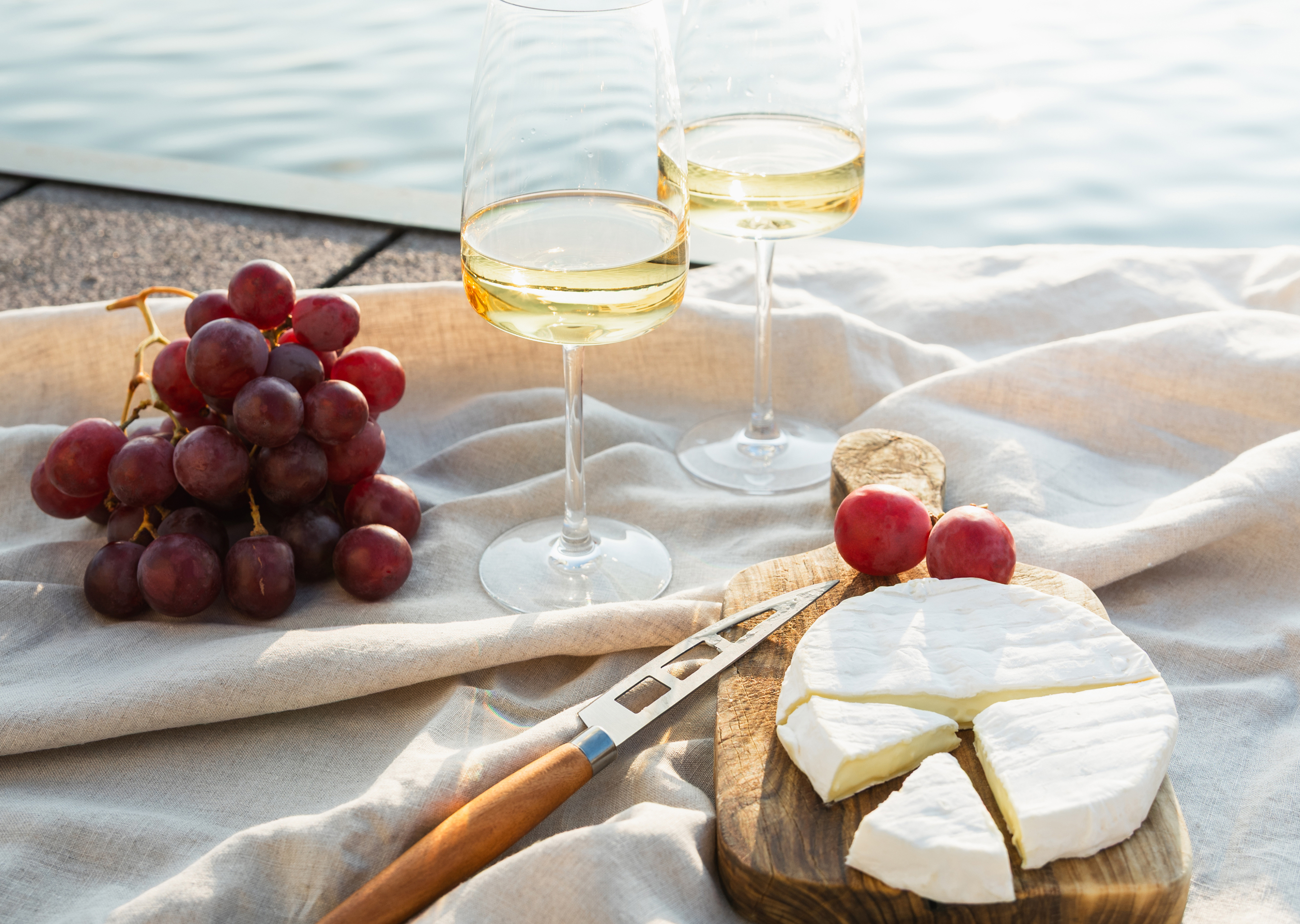 Zwei Gläser Secco mit Trauben und Käse auf einem Tuch am Wasser